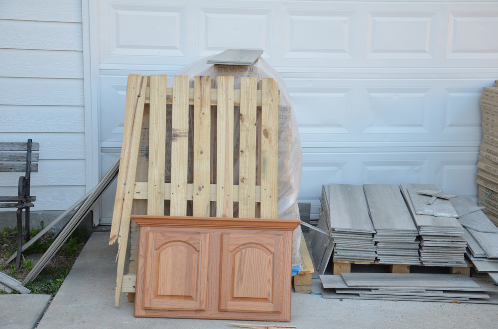 A wooden pallet and a pair of cabinet doors lean against a garage door, surrounded by stacks of tiles and construction materials.