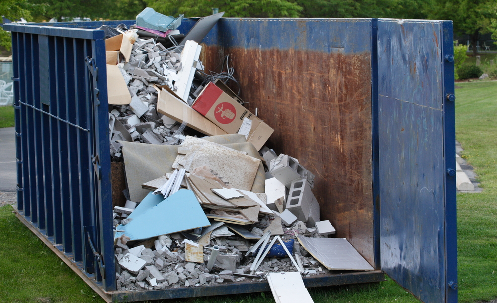 A large blue dumpster filled with construction debris, including broken drywall, concrete blocks, wood, and various building materials, sits on a grassy area beside a paved surface.