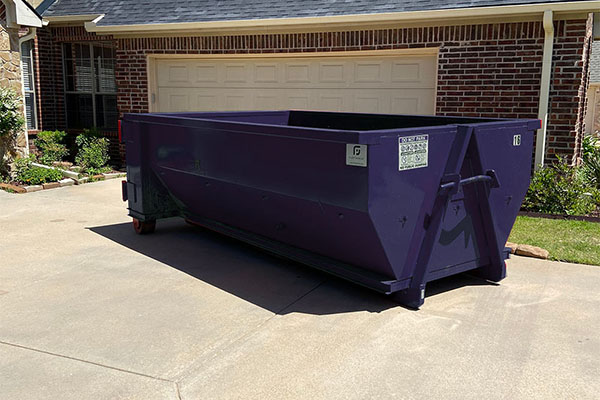 A truck parked on a gravel surface near a partially constructed house with wooden framing. Two large red dumpsters sit beside it. Trees and a clear blue sky are in the background.