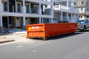 An orange dumpster labeled RENT ME sits on the street in front of houses under construction, next to a parked silver pickup truck. Yellow caution tape is visible near the entrance of one house.