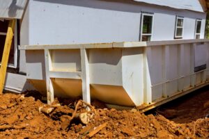 A large white metal dumpster sits on reddish dirt near a building under construction, with scattered debris and construction materials visible on the ground.