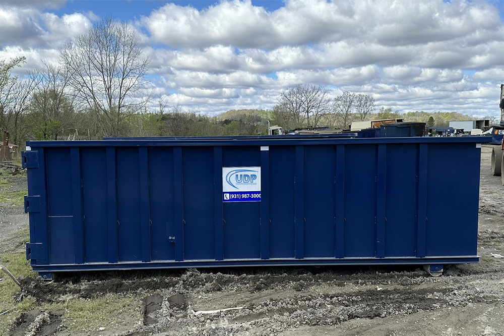 A large, rusty red dumpster filled with various debris, including cardboard, wood, black garbage bags, and other discarded materials, sits on a street near landscaped planters with green plants.